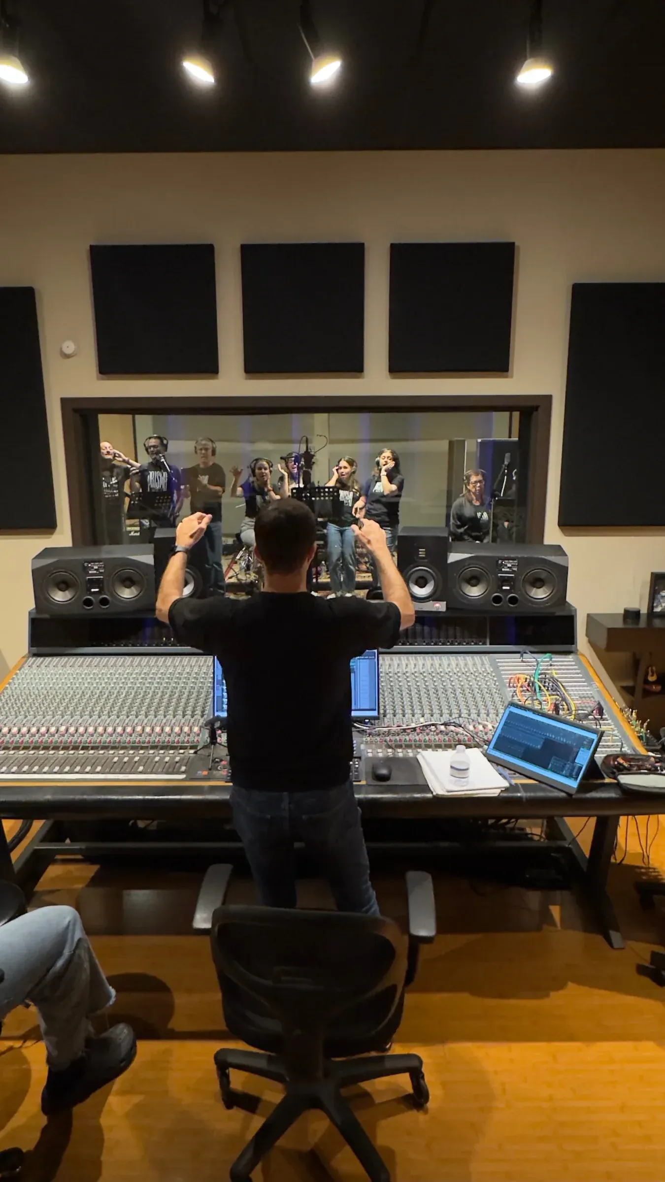 An engineer at the mixing desk, the group singing behind the studio glass